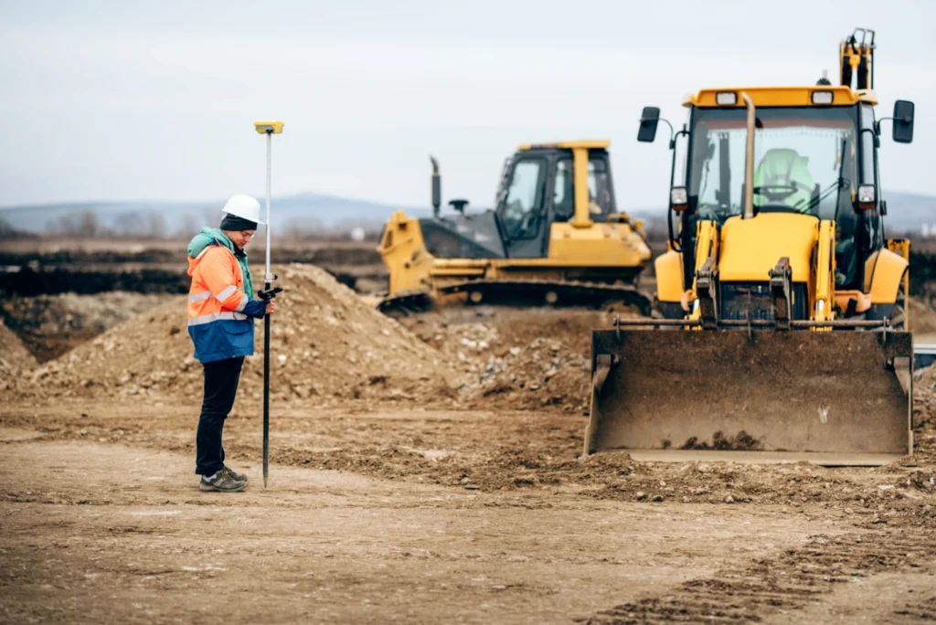 Construction worker using GPS surveying equipment on a job site with heavy machinery, including a bulldozer and backhoe, in the background. The image highlights the integration of modern technology in construction for precise grading and site preparation.