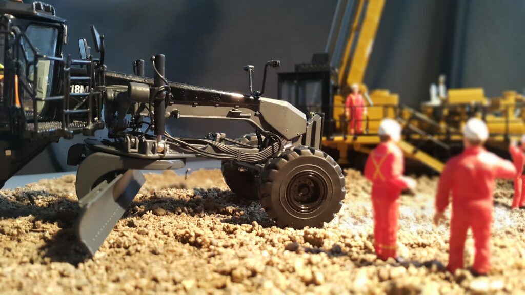 Close-up of a motor grader on a construction site, showcasing its detailed machinery components. In the background, construction workers and heavy equipment emphasize teamwork and the use of advanced tools for site preparation and grading while using material and earthwork takeoffs.